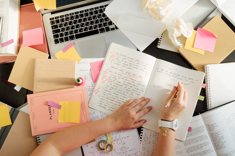 A student writing in a notebook with a laptop and planning materials nearby